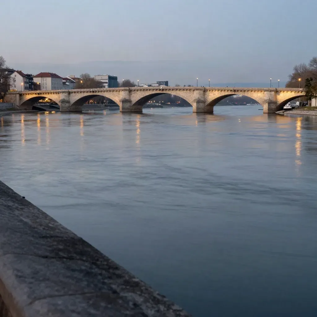Danube River at blue hour with bridge arches, lit riverfront and calm misty water.